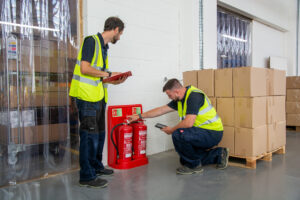 two engineers in a warehouse, wearing hi-vis jackets checking the quality of two extinguishers